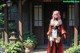 A woman in a red and black outfit stands outside a house with greenery around.