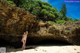 A naked woman standing in front of a cave on a beach.